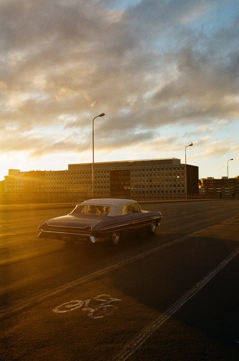A vintage convertible car drives on a road with a bicycle lane symbol painted on it, under a cloudy sky during golden hour.