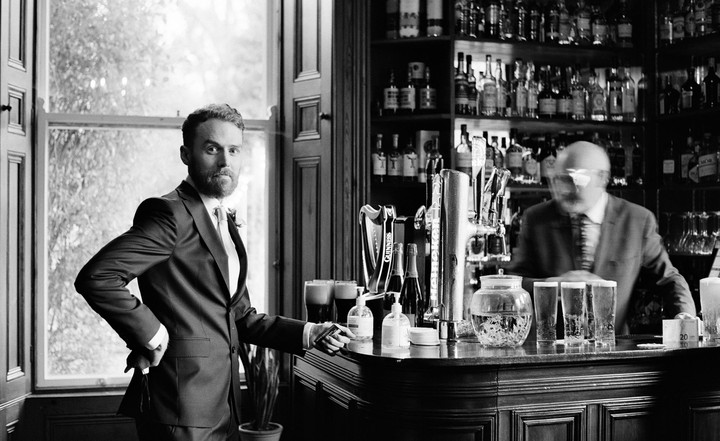A man in a suit stands by a window next to a bar with a blurred bartender in the background.
