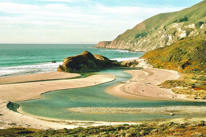A river flows into the ocean at a sandy beach with mountains in the background.