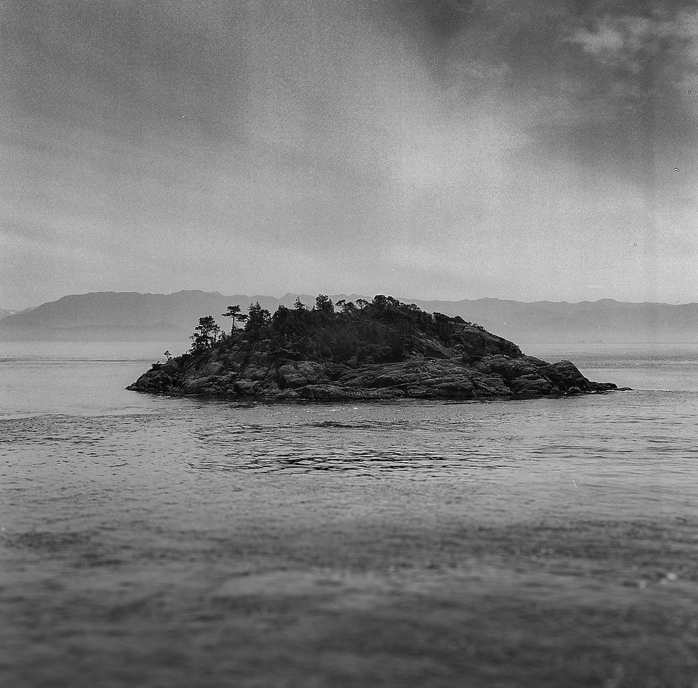 A small rocky island covered with trees sits in the water with mountains in the background under a cloudy sky.