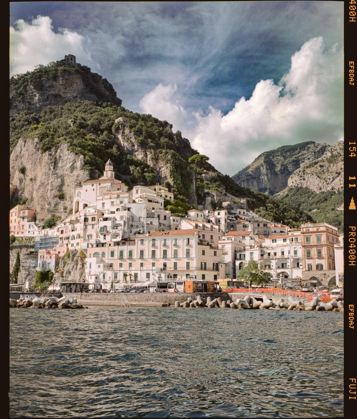 A coastal town with white buildings is nestled at the base of a large, green mountain, with water in the foreground.