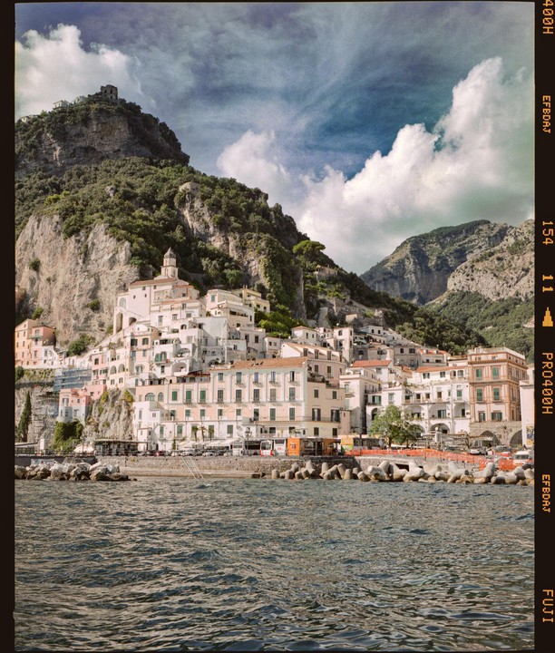 A coastal town with white buildings is nestled at the base of a large, green mountain, with water in the foreground.