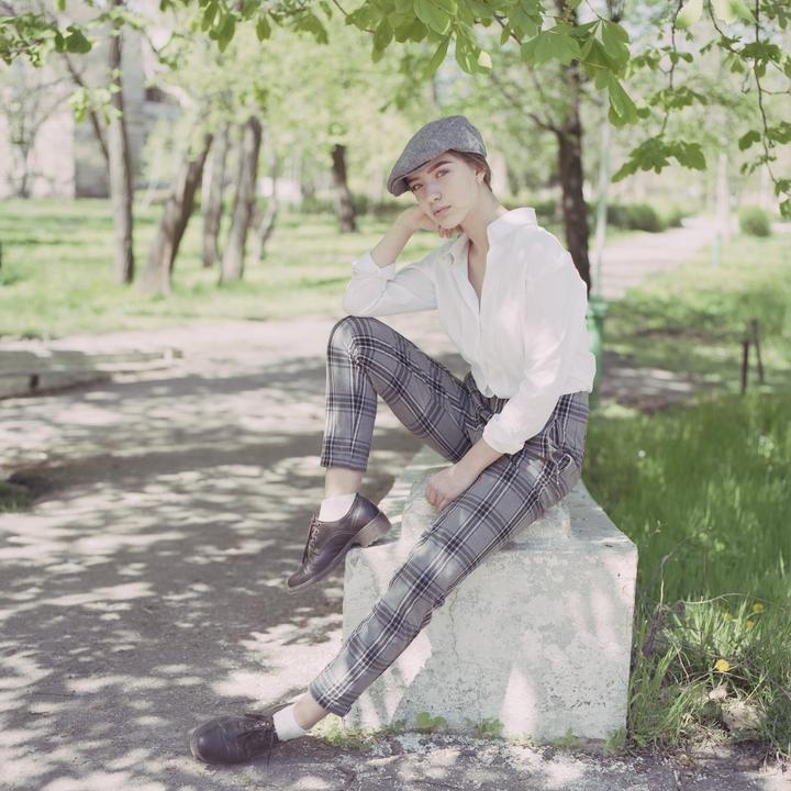 A woman wearing a flat cap and plaid trousers sits on a concrete block in a park.