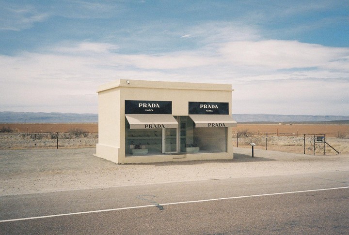 A small, standalone Prada store with awnings stands in a desert landscape next to a road.