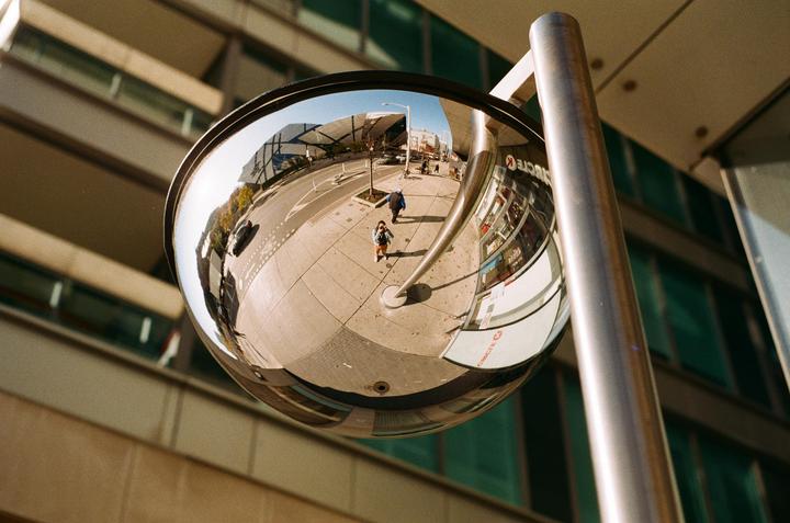 A convex mirror reflects a street scene with a woman and a man walking on a sidewalk.