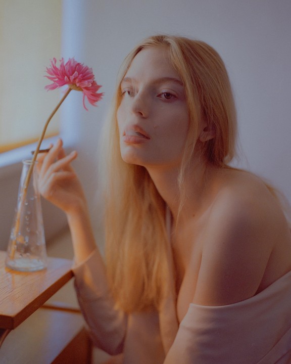 A woman with long blonde hair holds a pink flower in a vase on a wooden table.