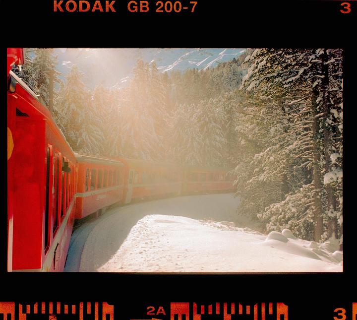A red train travels through a snowy forest with sunlight shining through the trees.