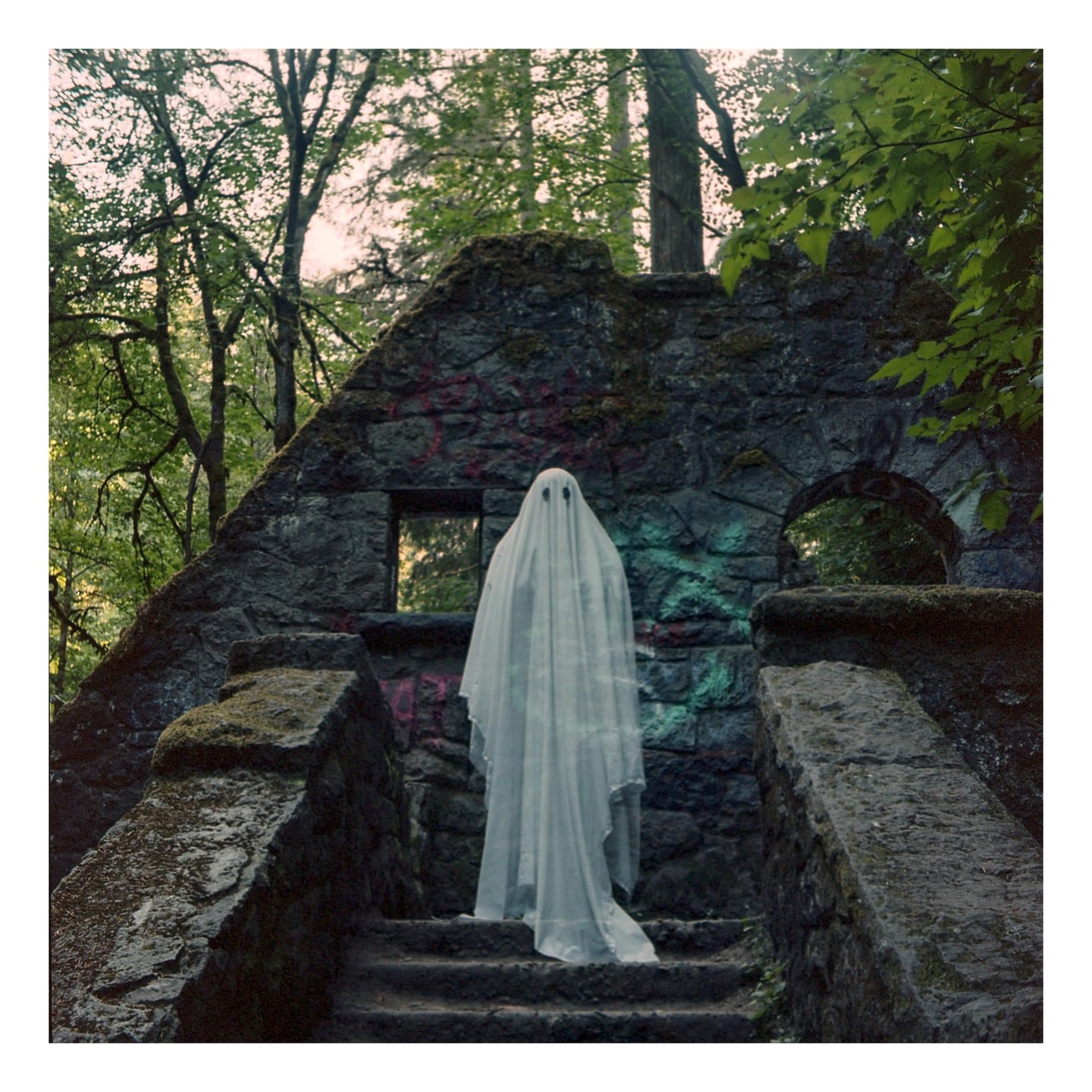 A person dressed as a ghost stands on stone steps in front of a ruined stone structure surrounded by trees.