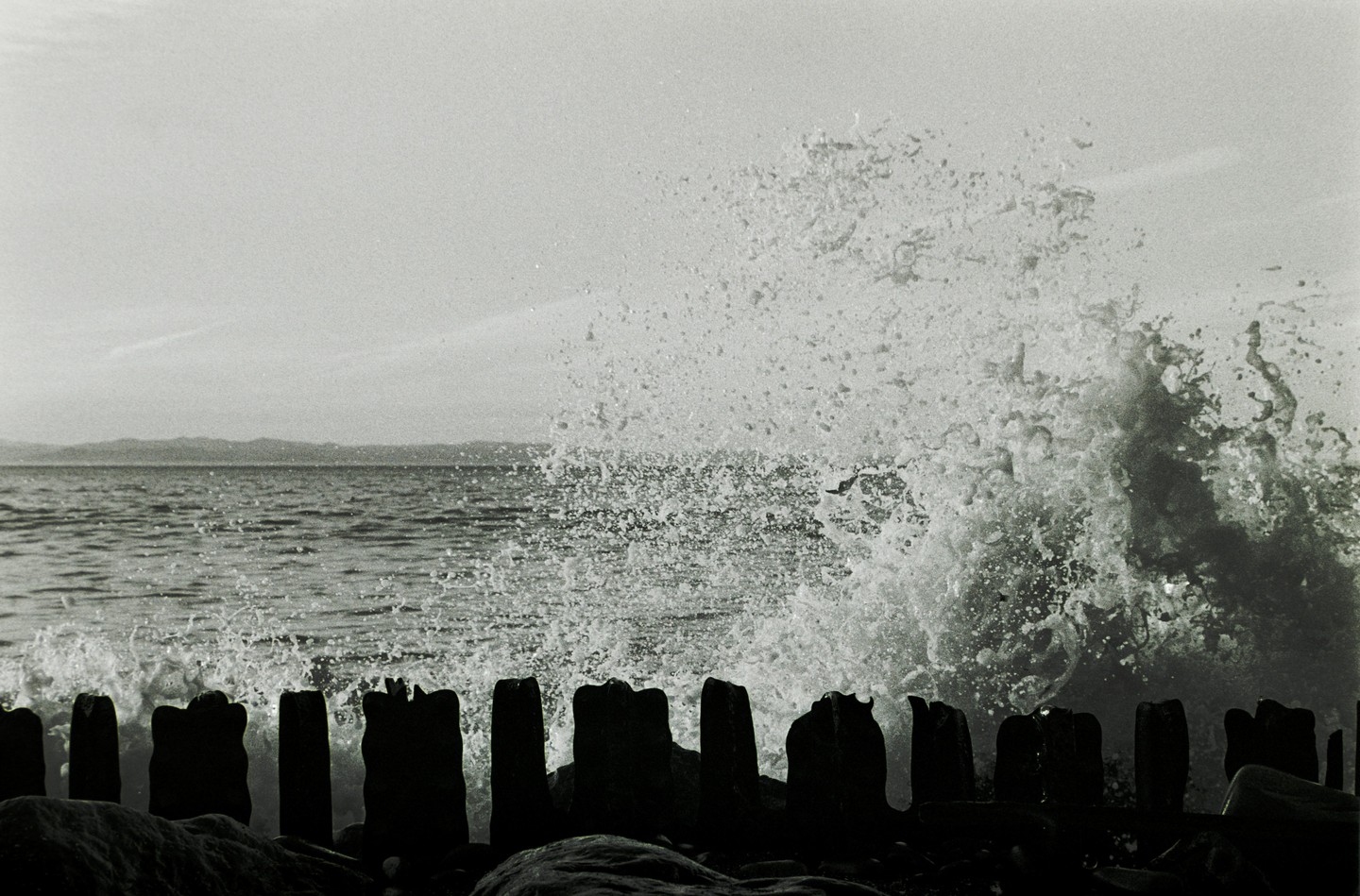 A large wave crashes over a wooden breakwater on a body of water with mountains in the distance.