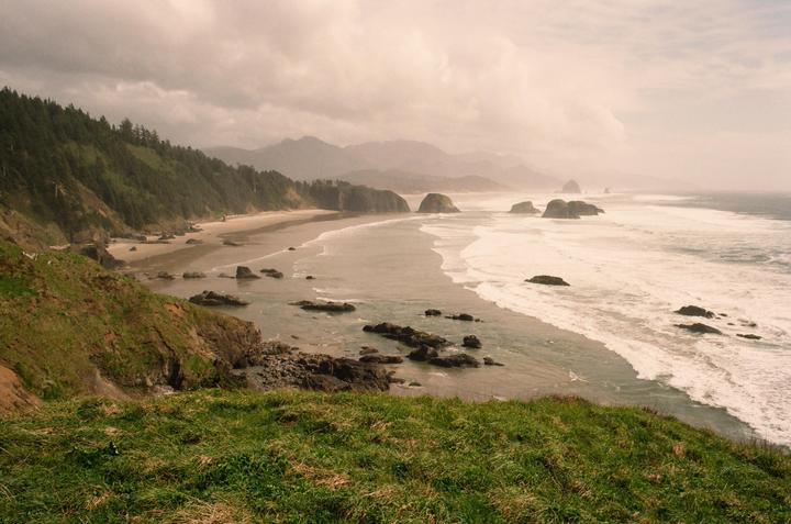 A wide view of the Oregon coast shows a sandy beach, forested hills, and rock formations in the ocean under a cloudy sky.