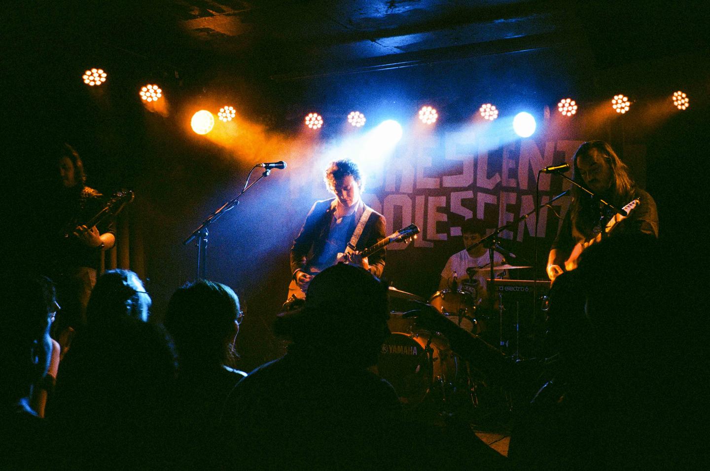A band performs on stage under bright orange and blue lights, with audience members visible in the foreground.