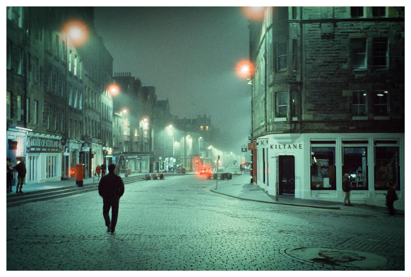 A man walks down a cobblestone street at night with buildings and streetlights on either side.