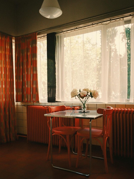 A small table with two chairs and a vase of flowers sits in front of a window with sheer white curtains and patterned orange curtains.