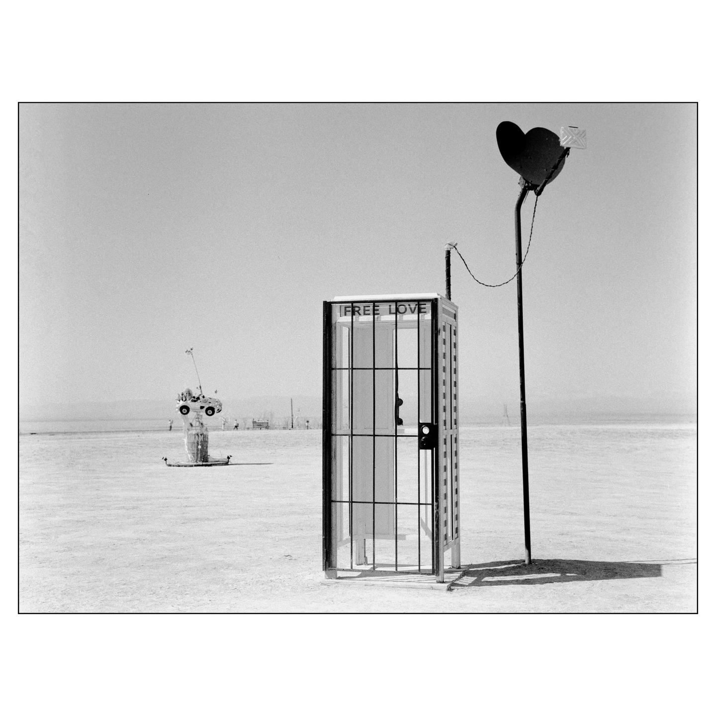 A telephone booth with a "FREE LOVE" sign stands in a desert landscape next to a pole with a heart-shaped sign.