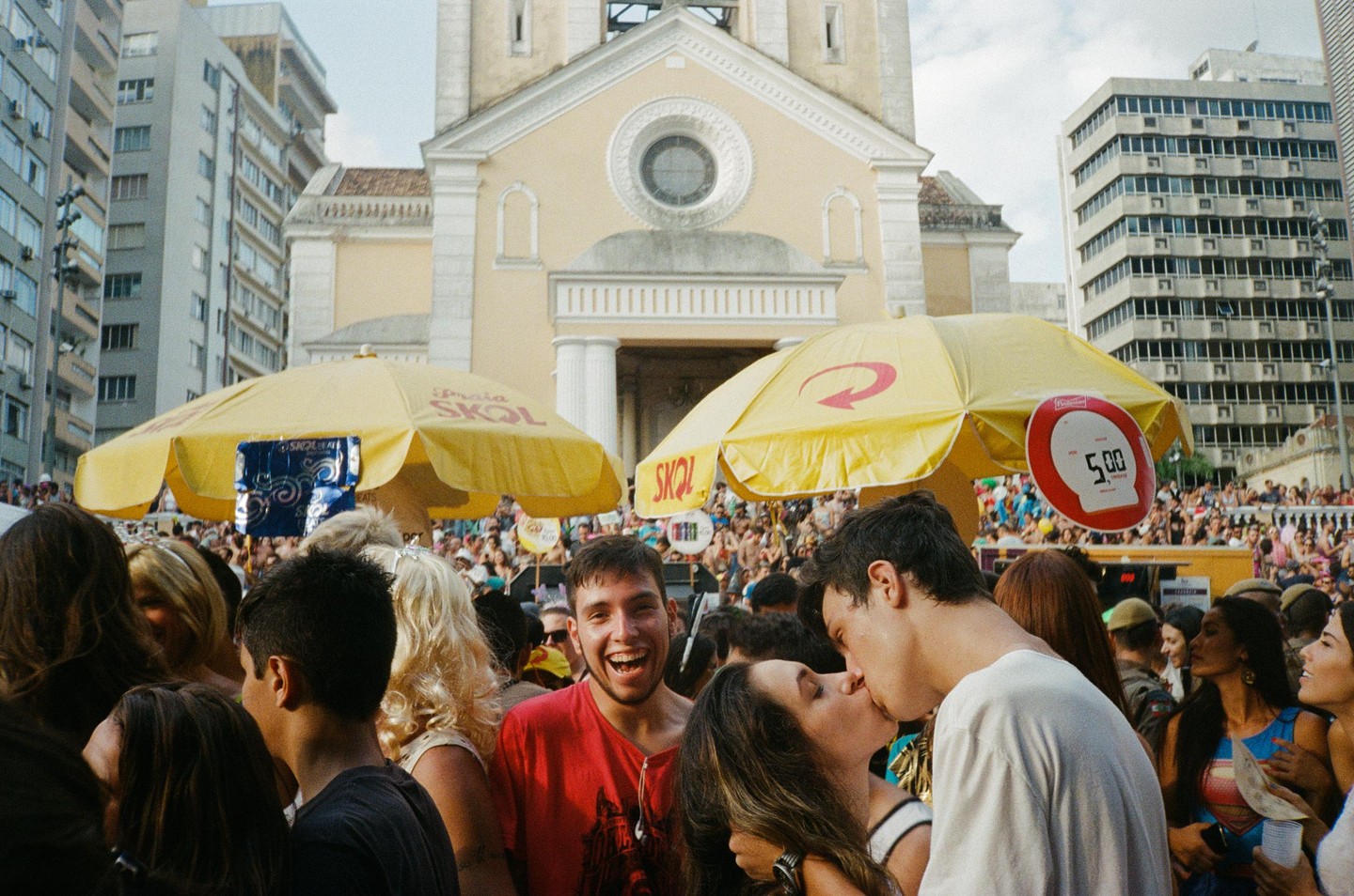 A couple kisses in a crowded street during Carnaval in Florianópolis, with a church and buildings in the background.