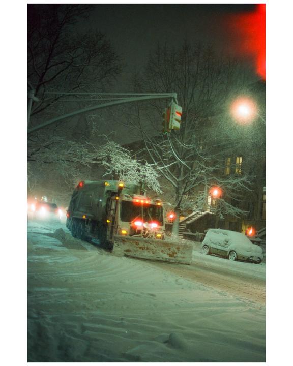 A snowplow clears a snow-covered street at night, with a car parked on the side of the road.