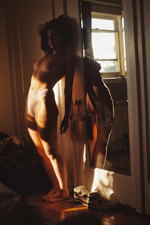 A woman stands topless in front of a mirror, illuminated by a golden light.