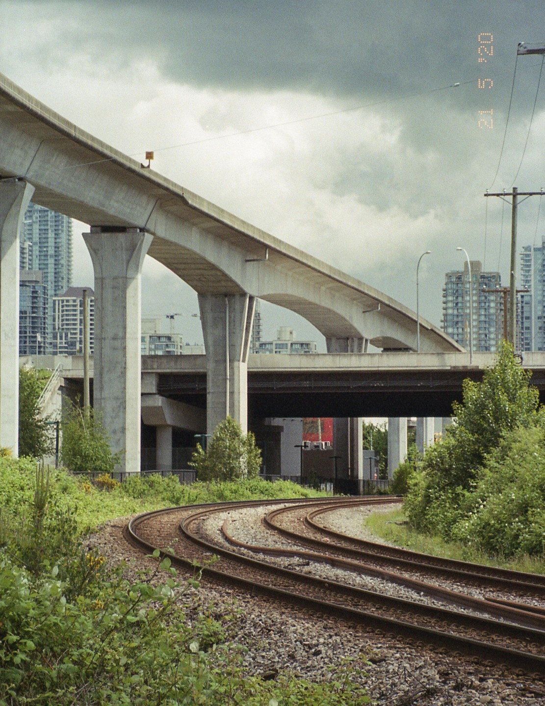A curved railway track runs through an urban landscape with elevated highways and buildings under a cloudy sky.