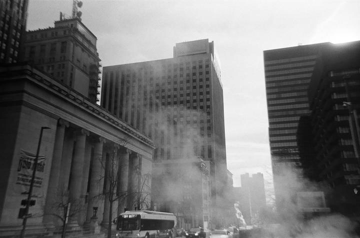A bus drives past a building with columns and several tall buildings in a hazy city.