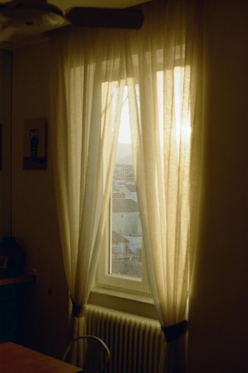 Sunlight streams through sheer curtains in a room with a view of rooftops.