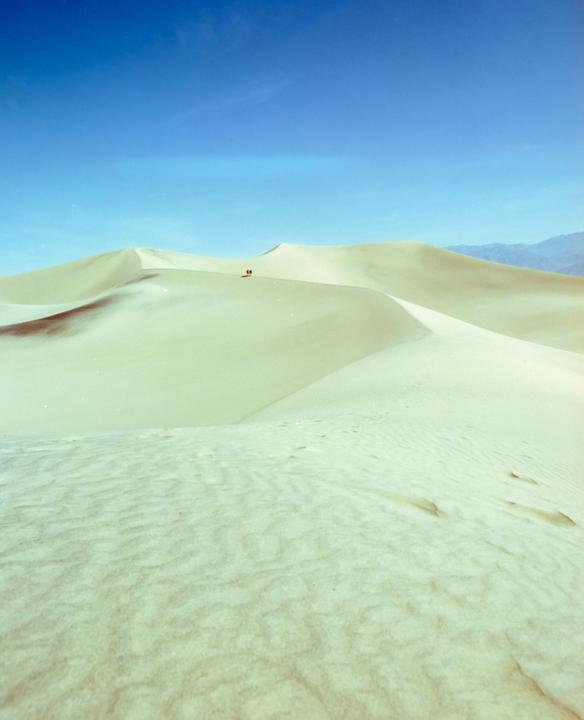 Two people walk across a vast desert landscape with sand dunes under a clear blue sky.