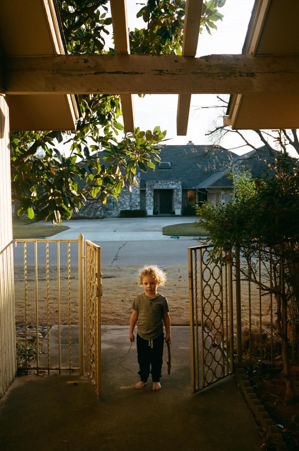 A child stands barefoot on a porch holding a stick, looking out through an open gate towards a house across the street.