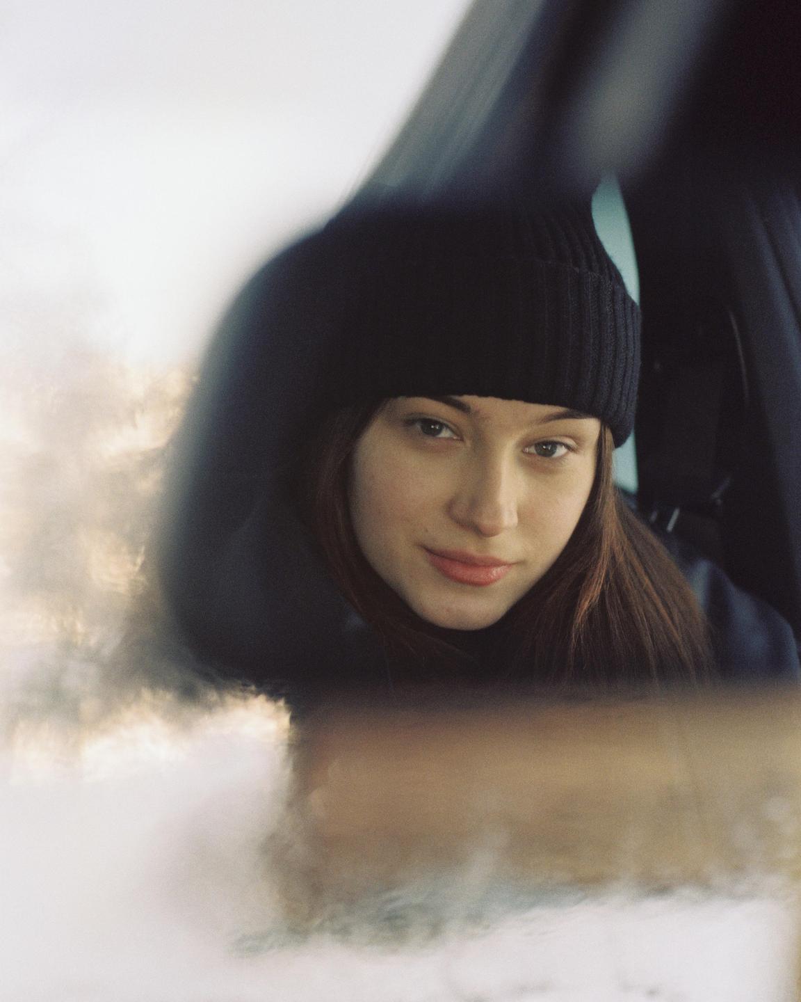 A woman wearing a black beanie looks directly at the viewer from inside a vehicle.