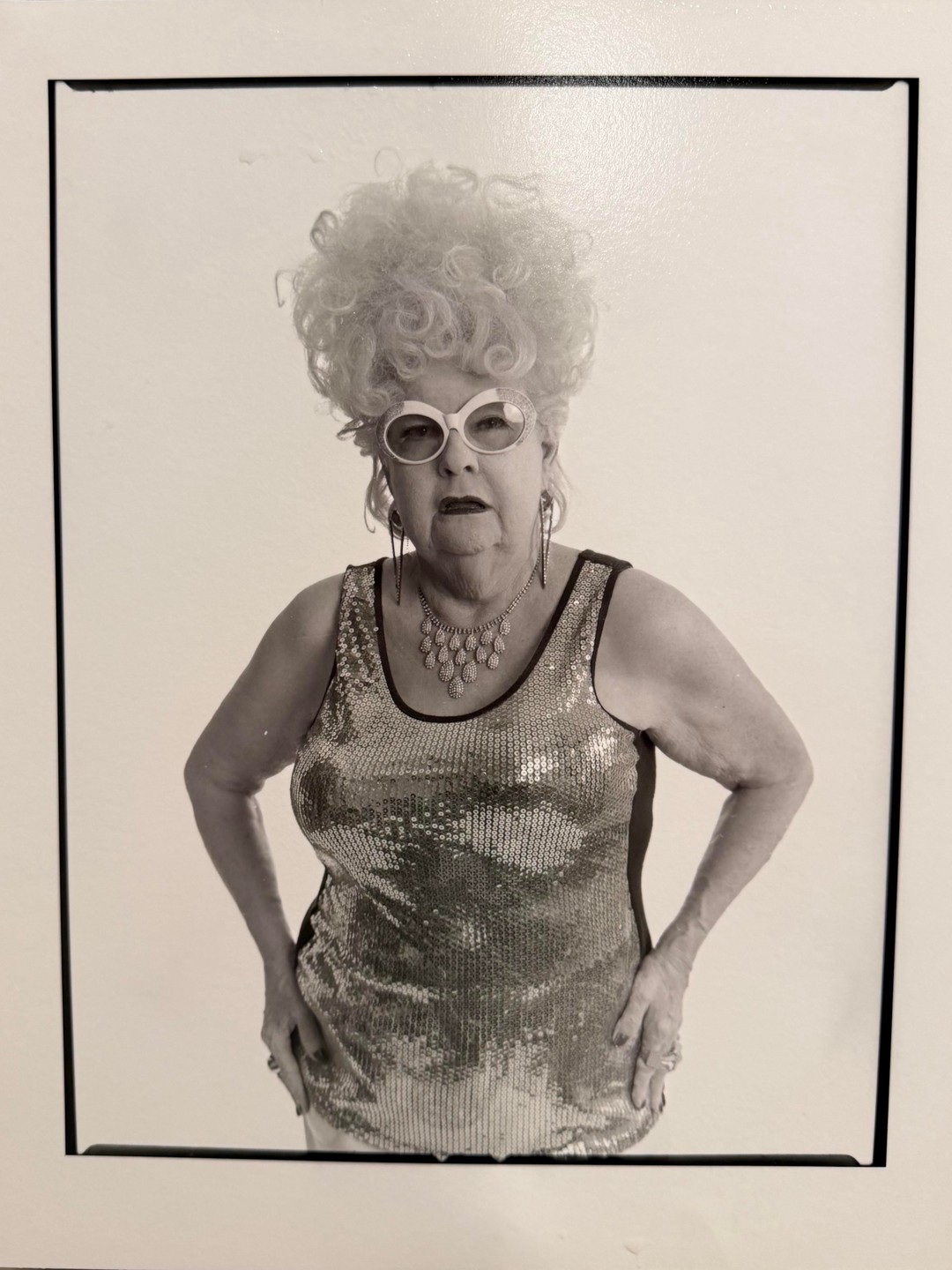 A woman with a large curly wig, sunglasses, and a sequined top poses with her hands on her hips.