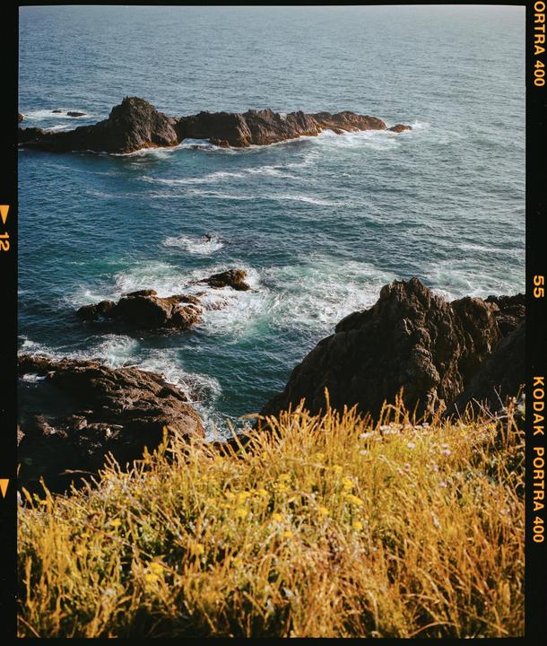 The Pacific Ocean crashes against a rocky coastline with a grassy cliff in the foreground.