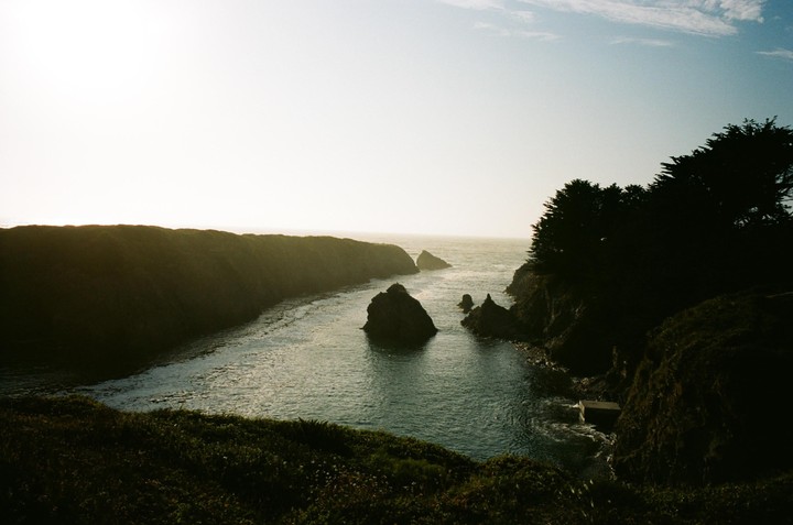 A body of water with several rock formations is surrounded by cliffs and trees under a bright sky.