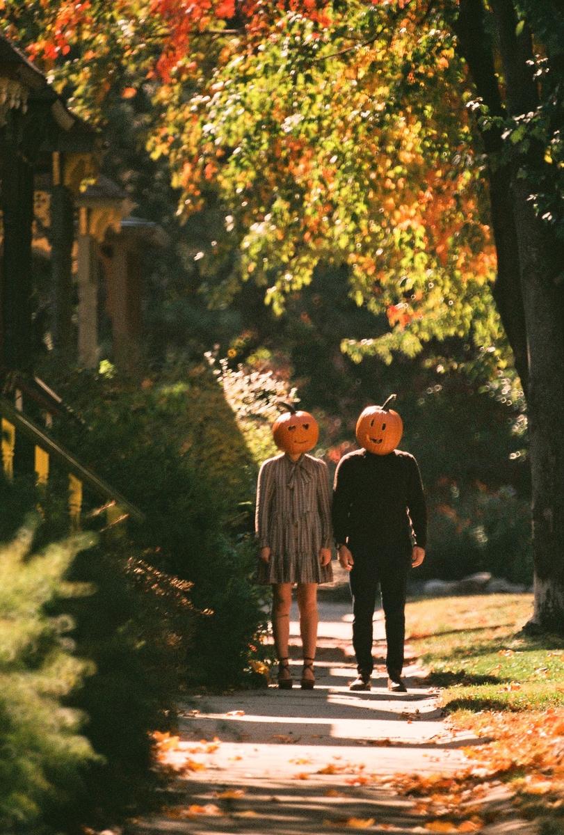 Two people with pumpkin heads stand on a sunlit path lined with autumn foliage.