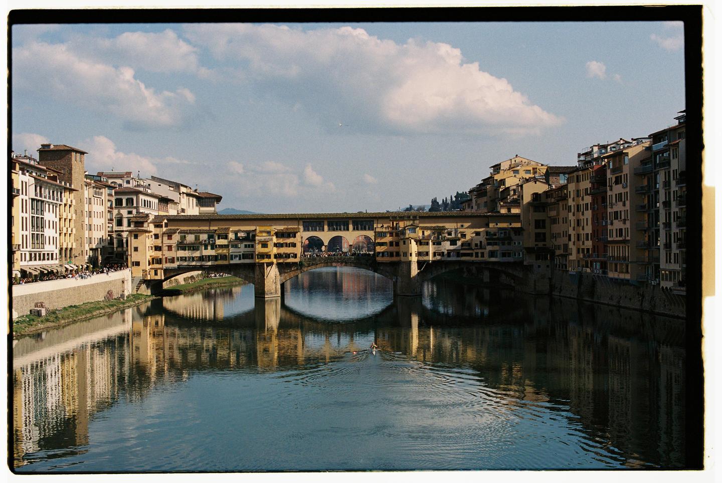 A bridge with buildings on top spans a river, with more buildings lining the riverbanks under a cloudy sky.