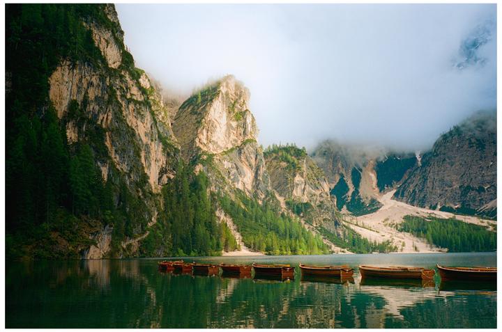 A line of wooden boats floats on a lake surrounded by mountains and forest.
