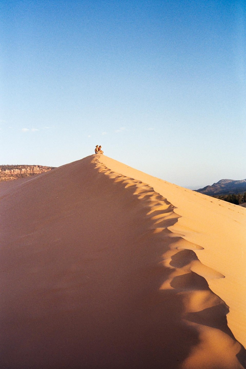 Two people sit atop a large sand dune under a clear blue sky.