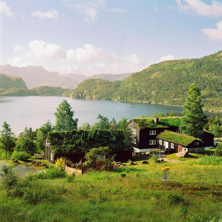 A cluster of houses with grass roofs sits on a hillside overlooking a lake surrounded by mountains.