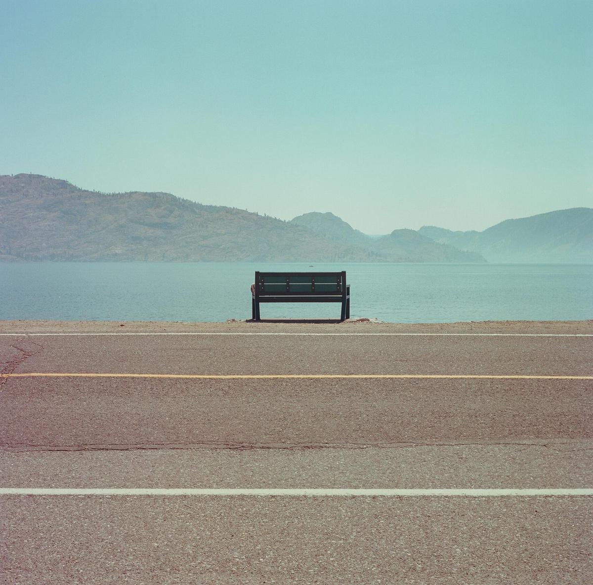 A lone bench sits beside a road overlooking a large body of water with mountains in the distance.