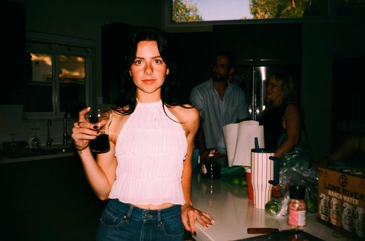 A woman holding a glass of red wine stands in a kitchen with other people in the background.