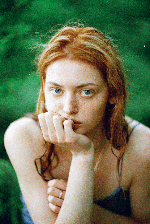 A woman with red hair and freckles looks directly at the viewer with her hand to her chin.