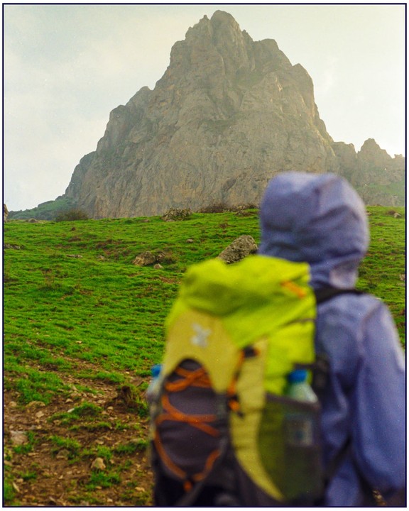 A person wearing a backpack and a hooded jacket stands on a grassy hill, looking up at a large mountain.