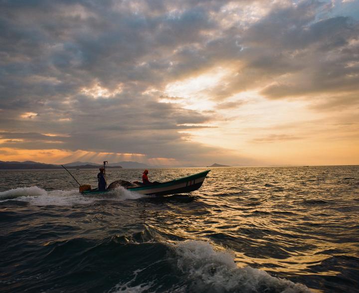 Two people are riding in a small boat on the water under a cloudy sky with sun rays breaking through.