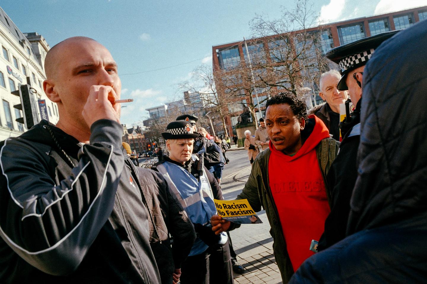A man in a red hoodie holds a "No Racism No Fascism" sign while talking to a police officer on a city street.