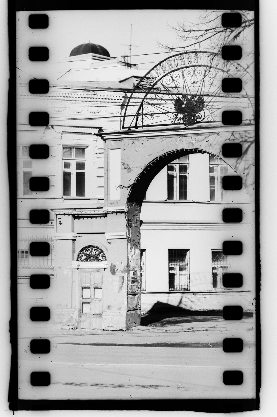 A black and white street photograph shows a building with an arched entrance and an ornate metal gate featuring a double-headed eagle.