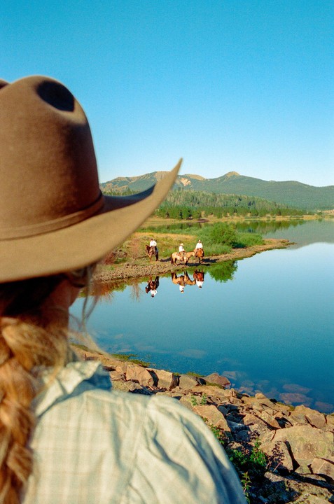 A person wearing a cowboy hat looks out over a lake at three people on horseback.