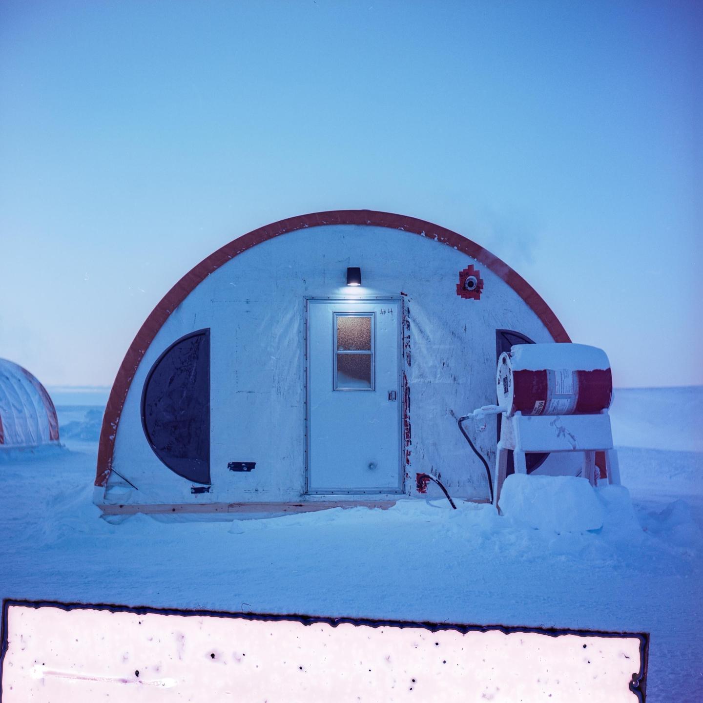 A small, white, arched building with an orange trim and a lit doorway stands in a snowy landscape under a clear sky.
