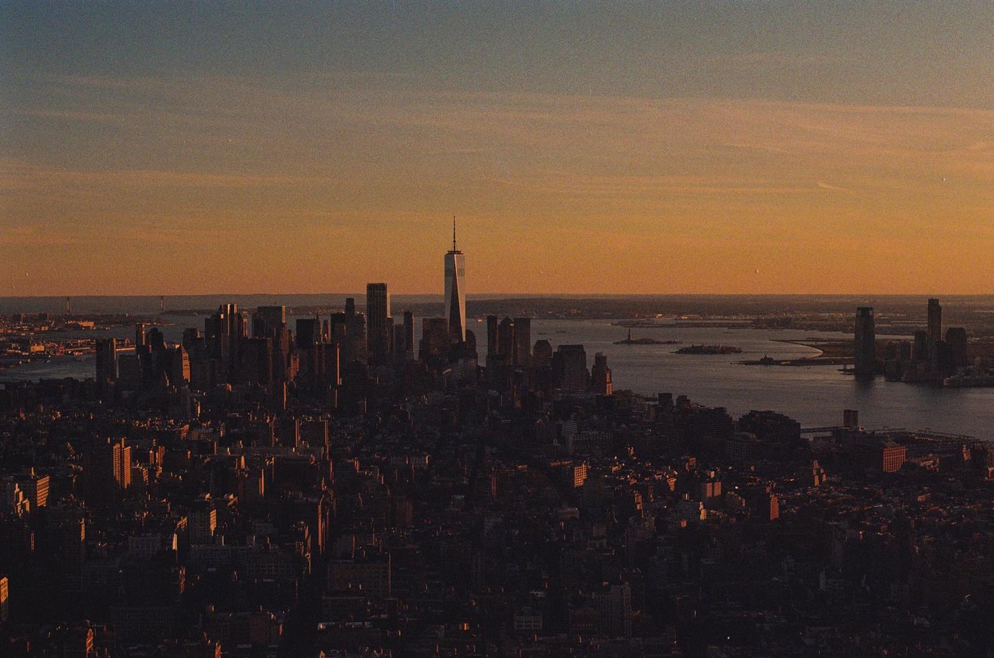 An aerial view of the New York City skyline at sunset shows the One World Trade Center building towering over other structures.
