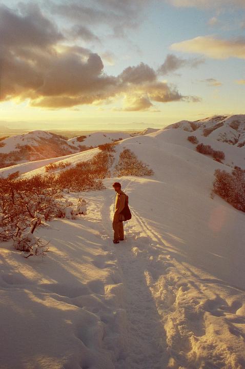 A man stands on a snowy mountain path at sunset, looking out over a valley.