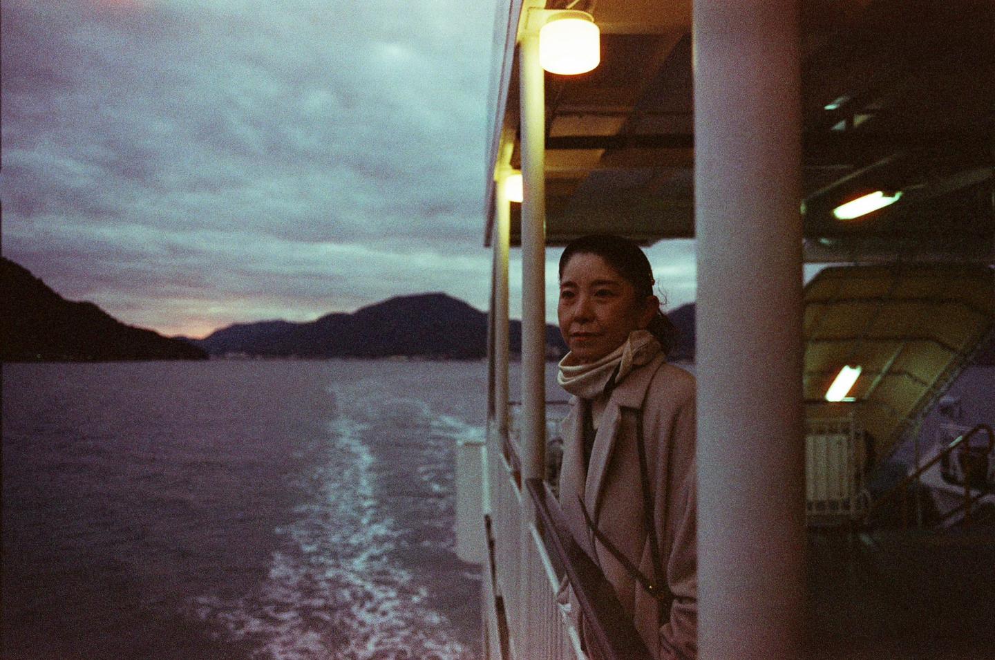 A woman stands on a boat deck at dusk, looking out at the water and mountains.