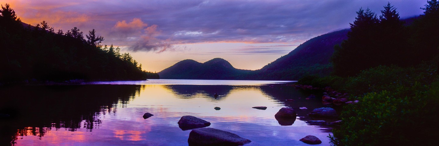 A panoramic landscape shows a lake surrounded by mountains and trees under a colorful sunset sky.