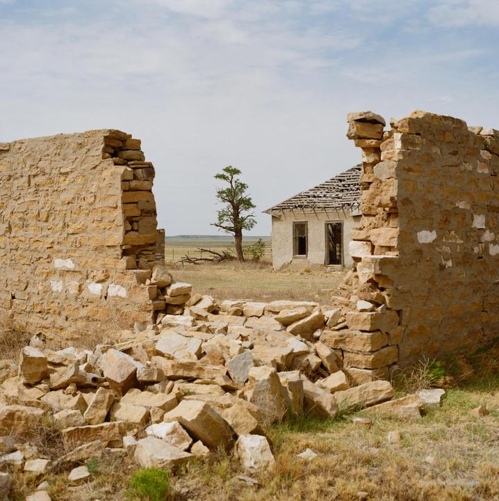 A dilapidated stone wall frames a view of a distant abandoned house and a lone tree in a dry, grassy field under a cloudy sky.
