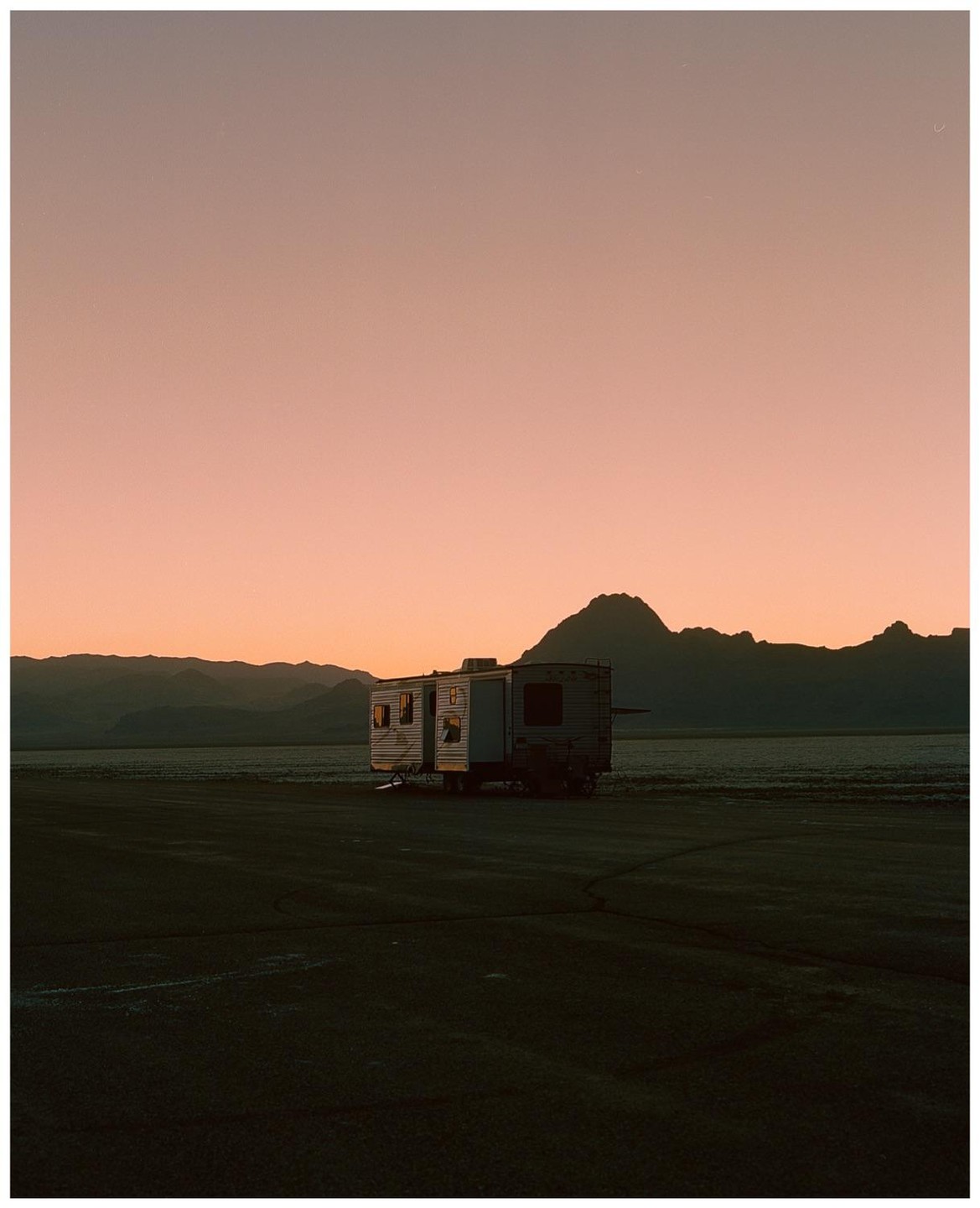 A camper trailer is parked in a vast, flat landscape with mountains in the background under a pink sky.